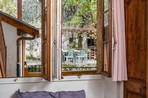 A room with a large window, wooden beams, and a view of outdoor furniture and greenery. - charming studio in the heart of the old town (Annecy)