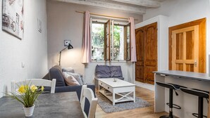 A compact living space with a sofa, a small table with a potted plant, a window with curtains, a wooden door, and a kitchenette area with bar stools. - charming studio in the heart of the old town (Annecy)