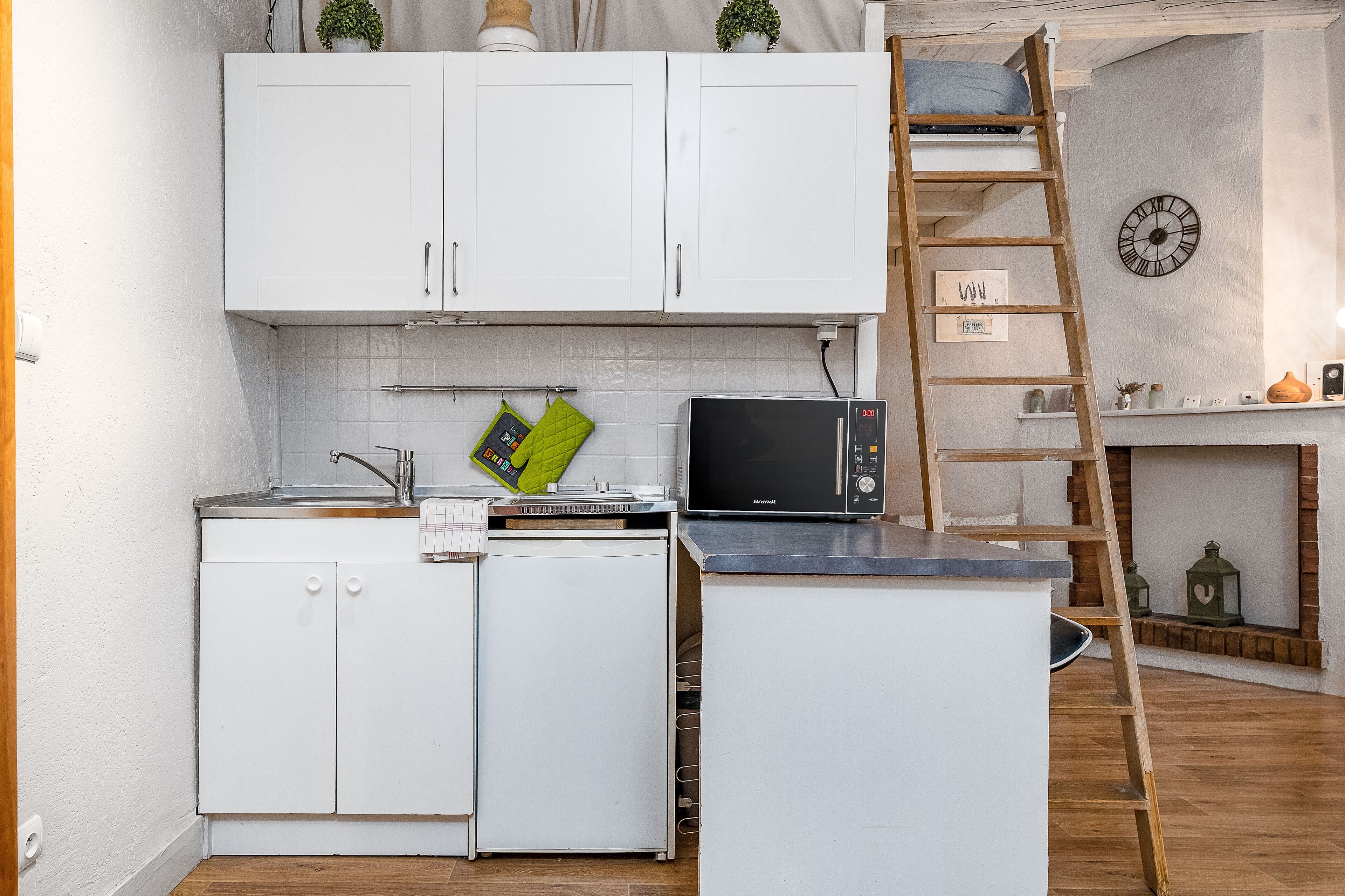 A compact kitchen with white cabinets, a microwave, and a small refrigerator. A wooden ladder is leaning against the wall, and a clock is visible on the wall.