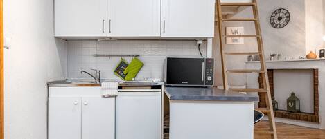 A compact kitchen with white cabinets, a microwave, and a small refrigerator. A wooden ladder is leaning against the wall, and a clock is visible on the wall.