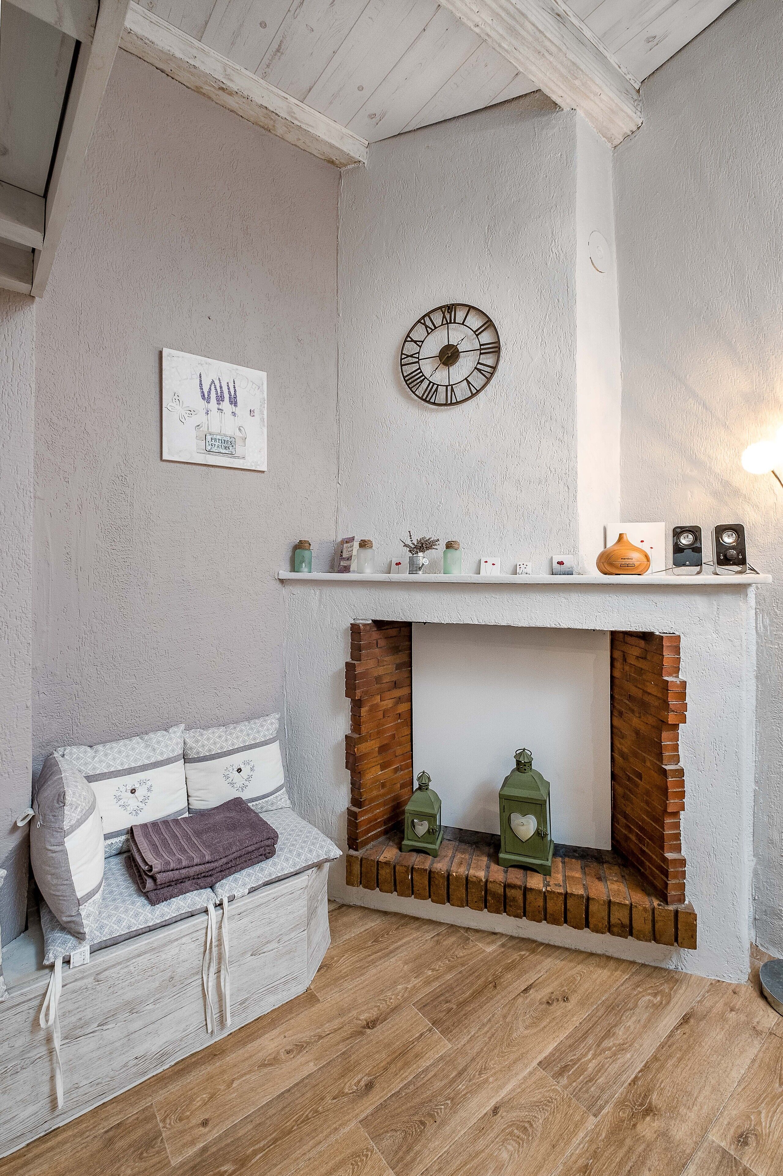 Une pièce chaleureuse avec un mur blanc, une horloge, une cheminée agrémentée d’objets décoratifs et un banc rembourré.