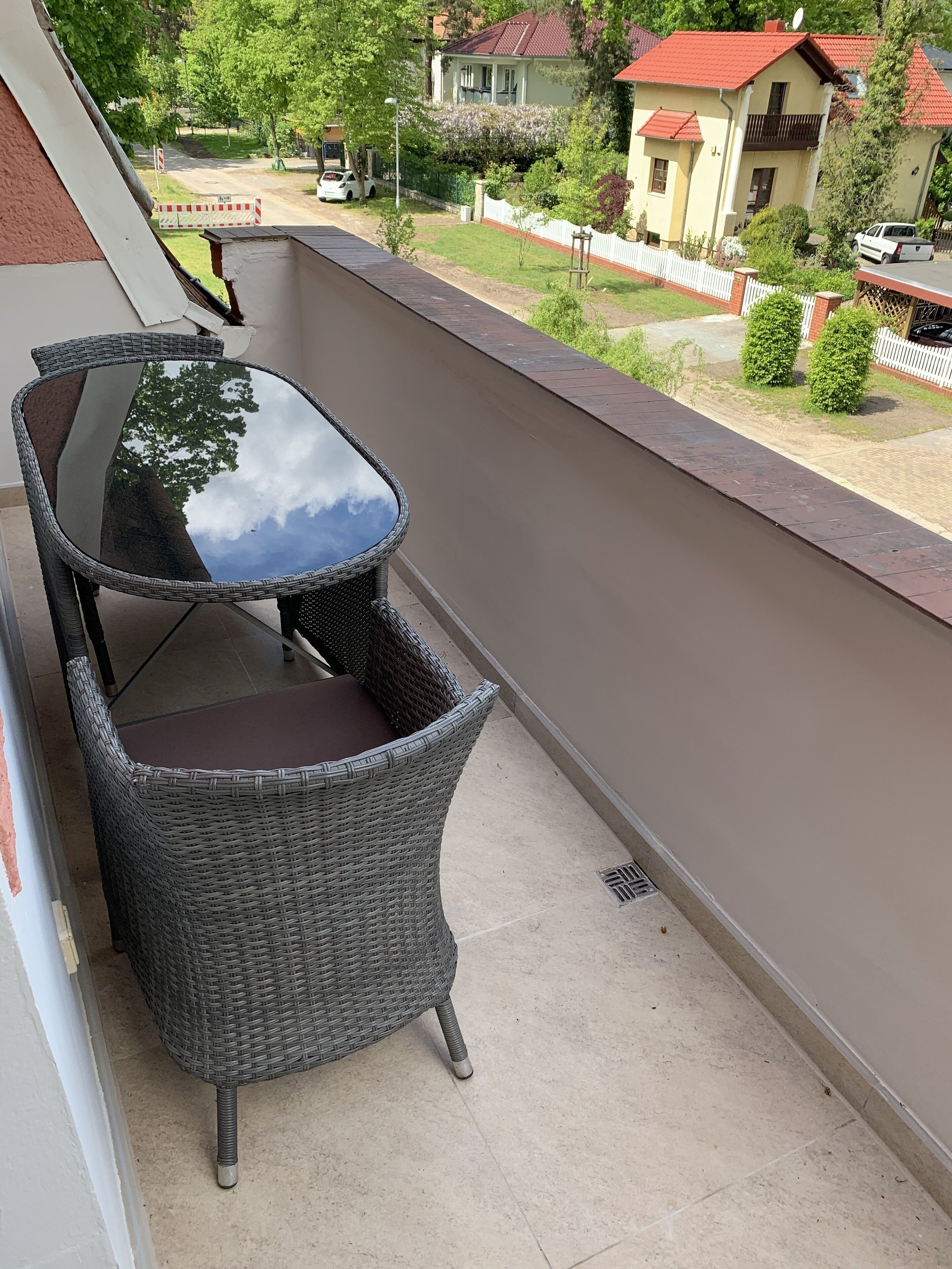 Un balcon avec un fauteuil en osier et une table en verre, donnant sur un quartier résidentiel avec des maisons et de la verdure.