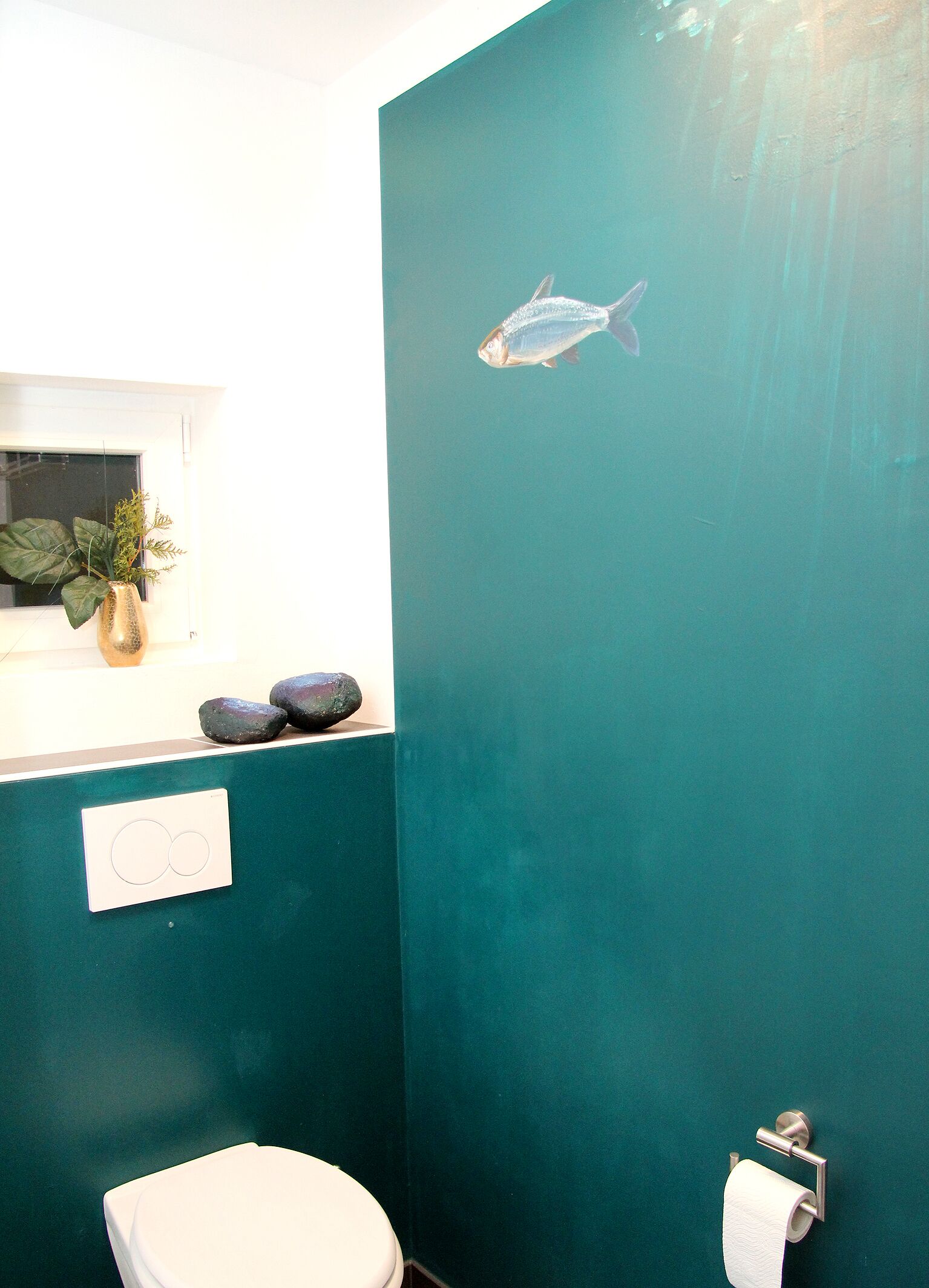 A bathroom with a teal wall, a toilet, a sink with a plant, and a fish decal.