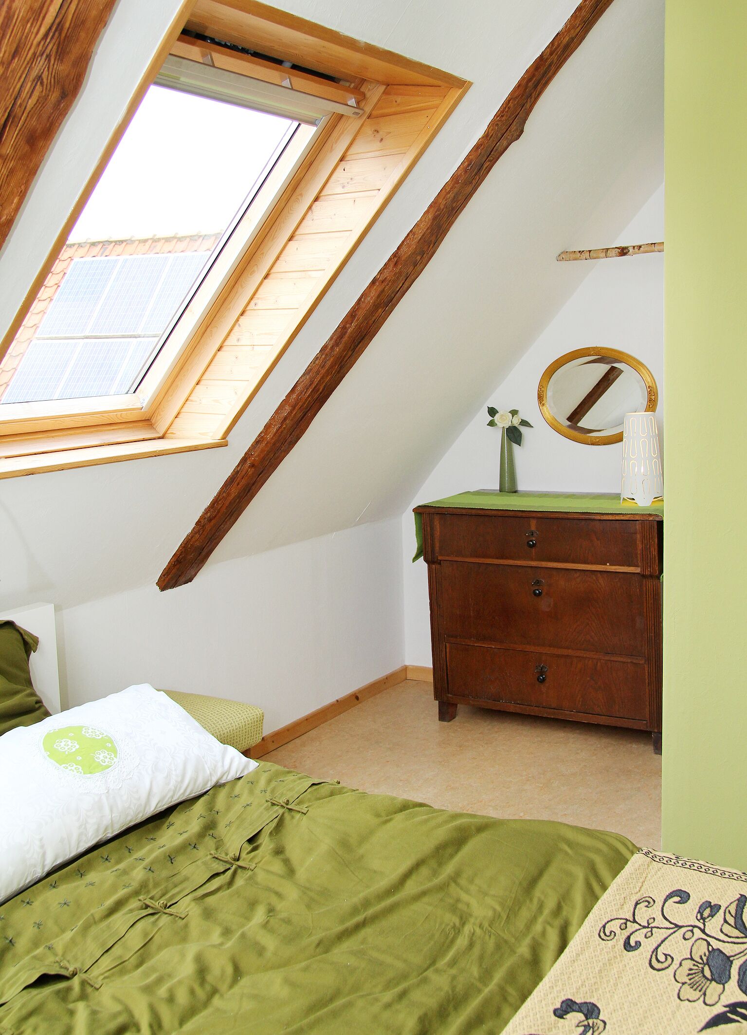 Un dormitorio con techo inclinado, tragaluz, cómoda de madera, espejo y cama con ropa de cama verde.