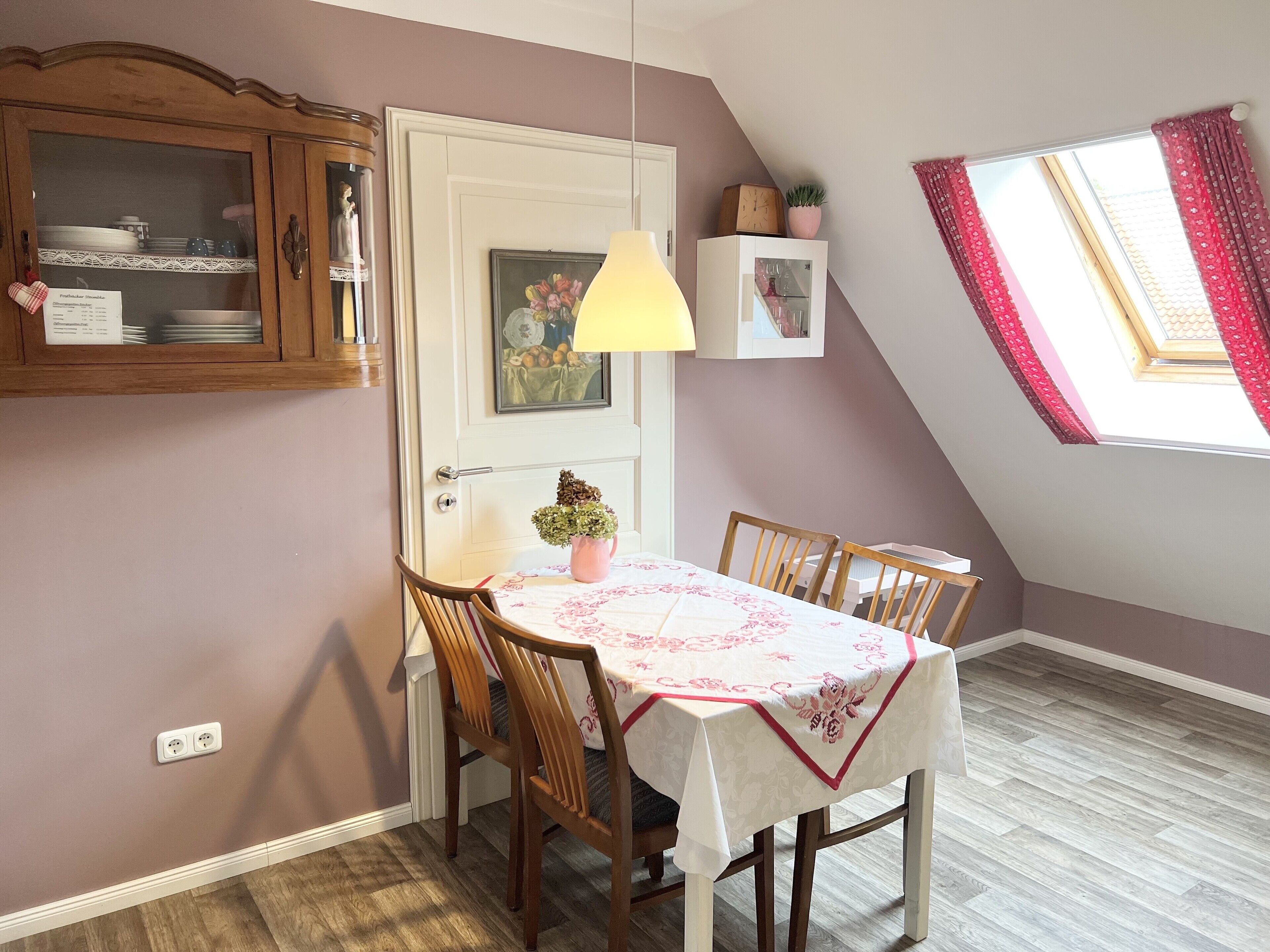 Un coin repas avec une table dressée pour quatre personnes, un buffet en bois avec des portes en verre, une porte ornée d’un tableau et une fenêtre avec des rideaux rouges.