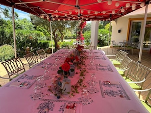 Un área de comedor al aire libre cubierta con una mesa larga preparada para una comida, rodeada de sillas y un toldo rojo.