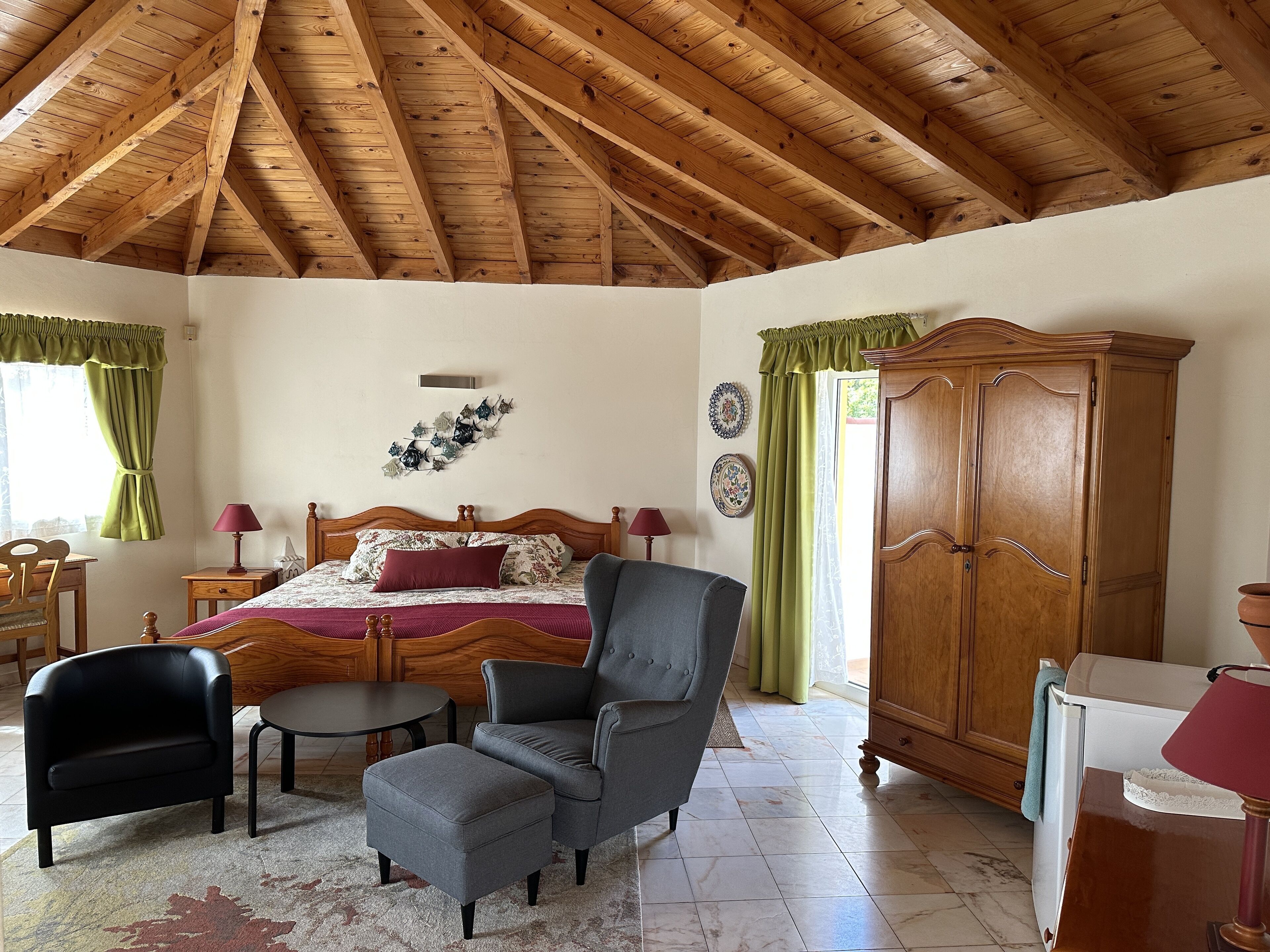 Ein Schlafzimmer mit Holzbalkendecke, einem Bett mit Blumenmuster-Bettwäsche, einem grauen Sessel, einem Holzschrank und einem kleinen Tisch.
