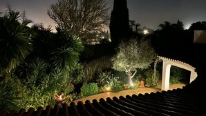 Een tuin bij nacht met een betegeld dak, een witte pergola en diverse planten.