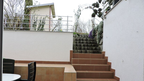 A patio area with a table and chairs, a set of stone steps leading to a higher level, and a white wall with a metal railing. - Away from the city life and yet part of the action (Appenweier)