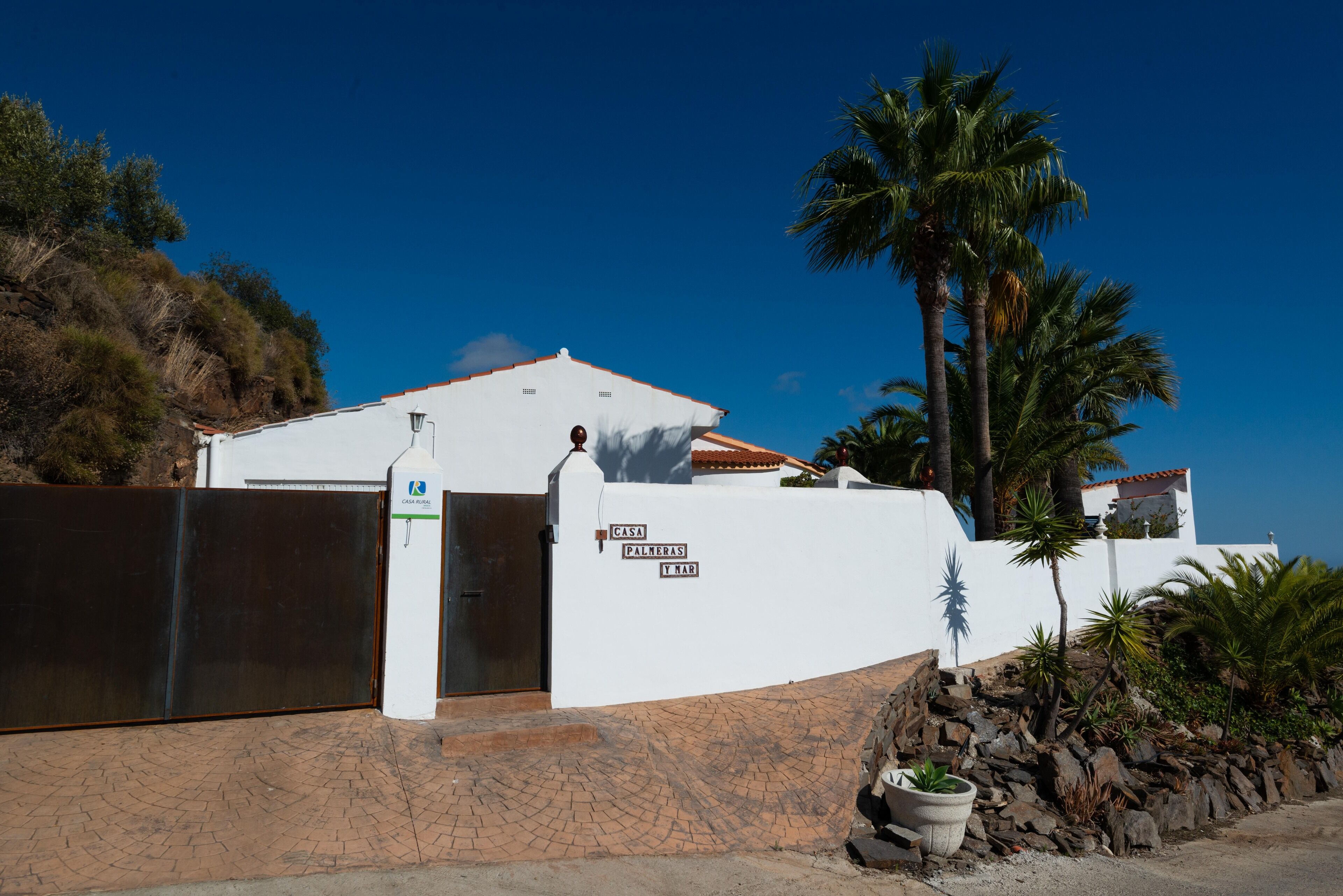 Ein weißes Gebäude mit einem befestigten Eingang, einem Schild mit der Aufschrift 'CASA PALMERAS Y MAR' und einer Palme im Hintergrund.