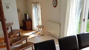 A dining area with a wooden table and chairs, a small side table, a radiator, a clock on the wall, and a view through a window.