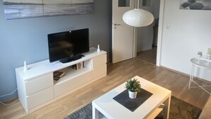 Una sala de estar moderna con un televisor de pantalla plana sobre una consola blanca, un tapete gris y una mesa de centro blanca con una planta.