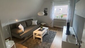 Una sala de estar moderna con un sof á gris, una mesa de centro blanca y un ventanal con cortinas.