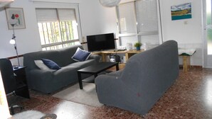 Un salon avec un canapé gris, une table basse, un téléviseur et un fauteuil. Il y a une fenêtre avec des stores, une lampe et un tableau au mur.