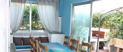 Un coin repas avec une table recouverte d’une nappe bleue, des chaises en bois et un banc agrémenté de coussins.