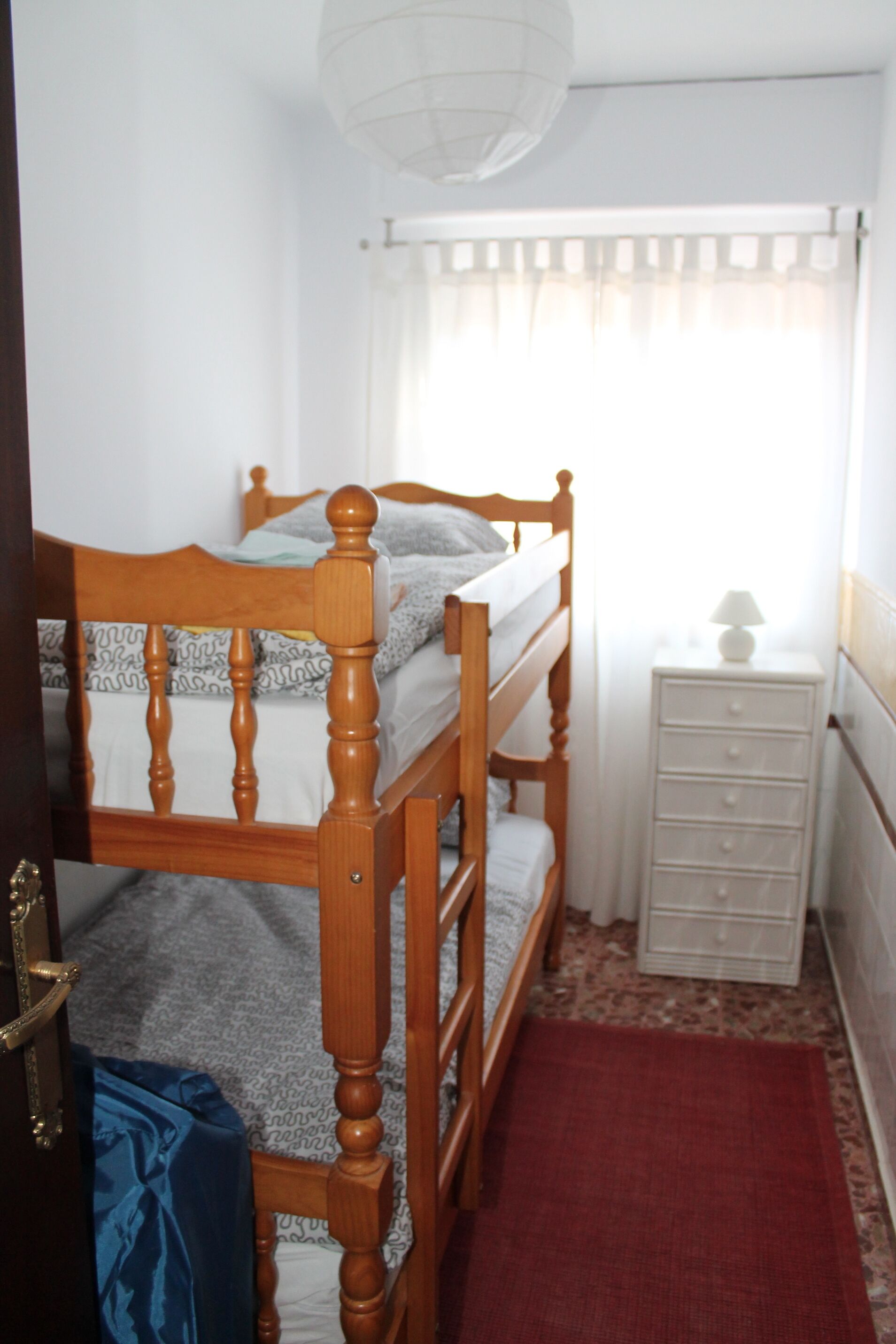 A bedroom with a wooden bunk bed, a white bedside table with a lamp, and a red rug.