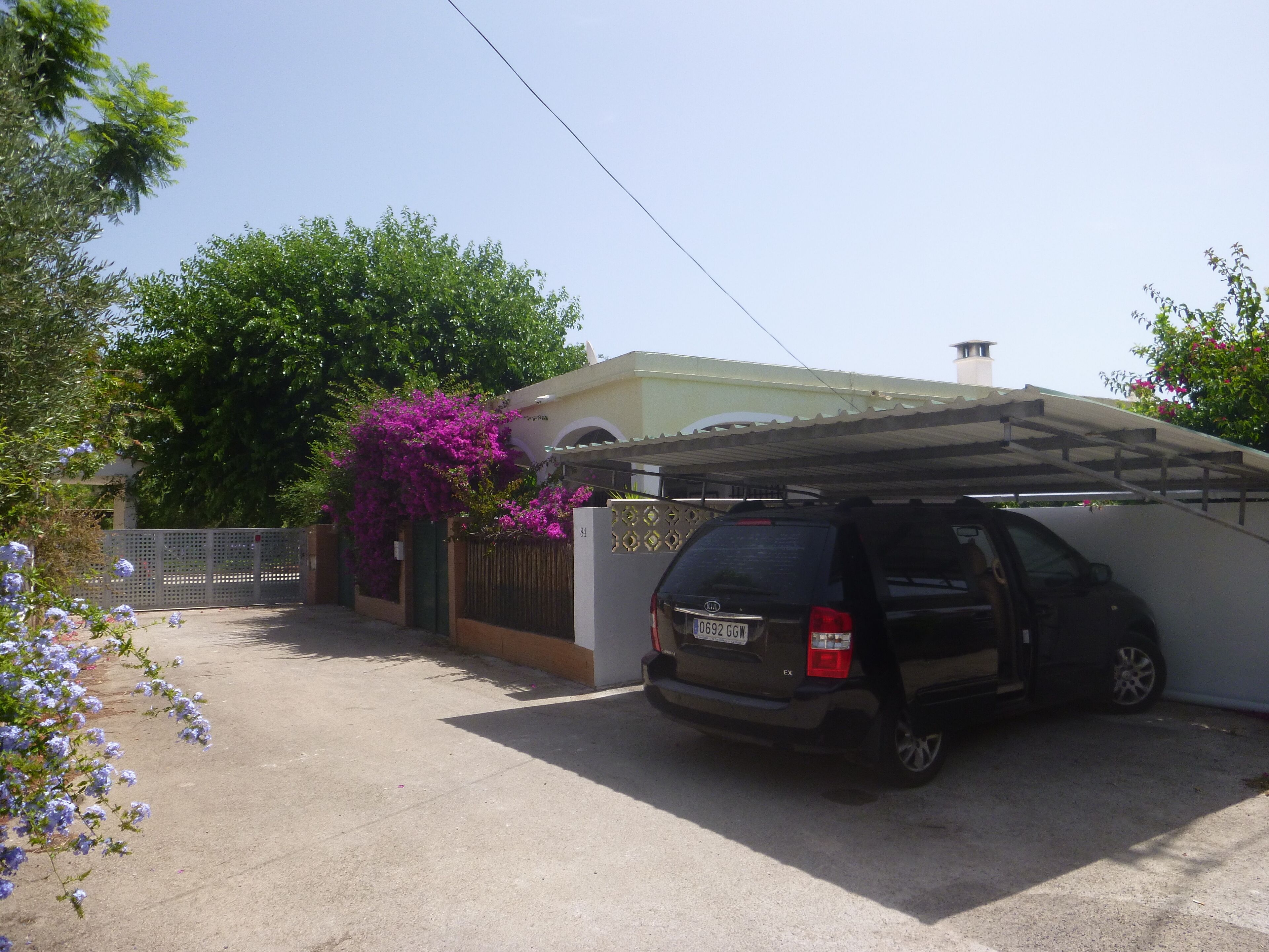 A black SUV parked in a driveway with a covered garage, a white fence, and purple bougainvillea flowers.