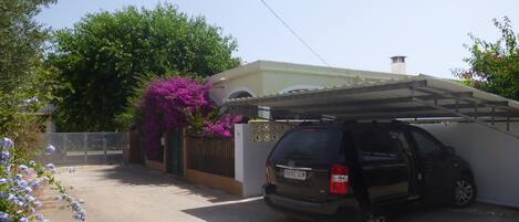 A black SUV parked in a driveway with a covered garage, a white fence, and purple bougainvillea flowers.