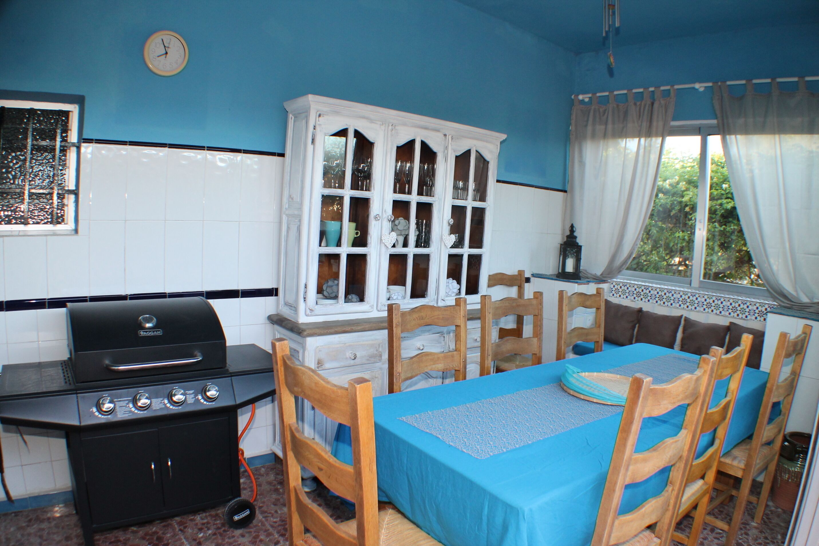 A kitchen with a grill, wooden chairs, a table with a blue tablecloth, and a cabinet with glass doors.