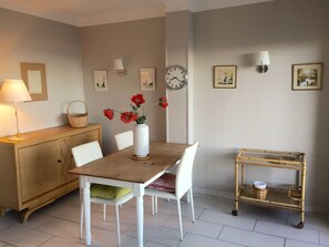 A dining area with a wooden table, white chairs, a vase with red flowers, a wall clock, and a small shelf with baskets. - Large 2 bedroom standing air conditioned seafront LINEN SUPPLIED / BED DONE (Canet-En-Roussillon)