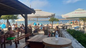 A beachside café with outdoor seating and umbrellas, overlooking a crowded beach and the sea.