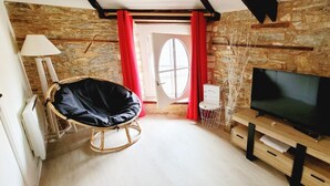 A cozy room with a stone wall, a black bean bag chair, a TV on a wooden stand, and a door with red curtains. - Charming Apartment - Downtown (Châteaubriant)
