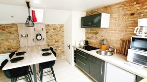 A compact kitchen with a stone wall, a wooden shelf with clocks, a dining table with chairs, and modern appliances. - Charming Apartment - Downtown (Châteaubriant)