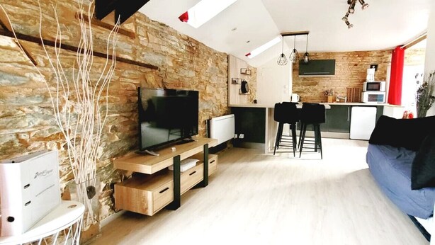 A modern living room with a stone wall, a flat-screen TV, wooden furniture, and a kitchen in the background. - Charming Apartment - Downtown (Châteaubriant)