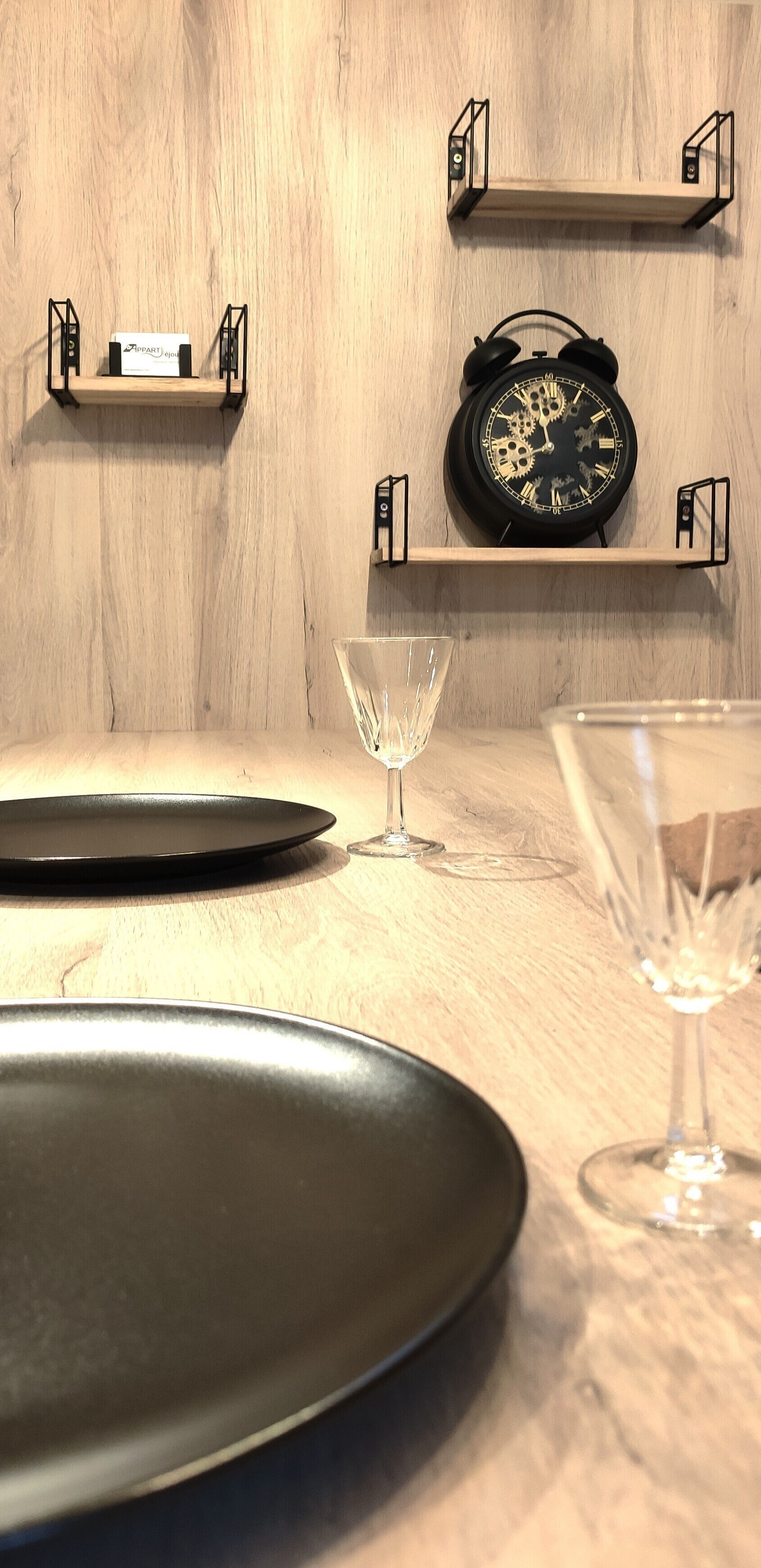 A wooden wall with two black shelves, one holding a clock and the other a small box.