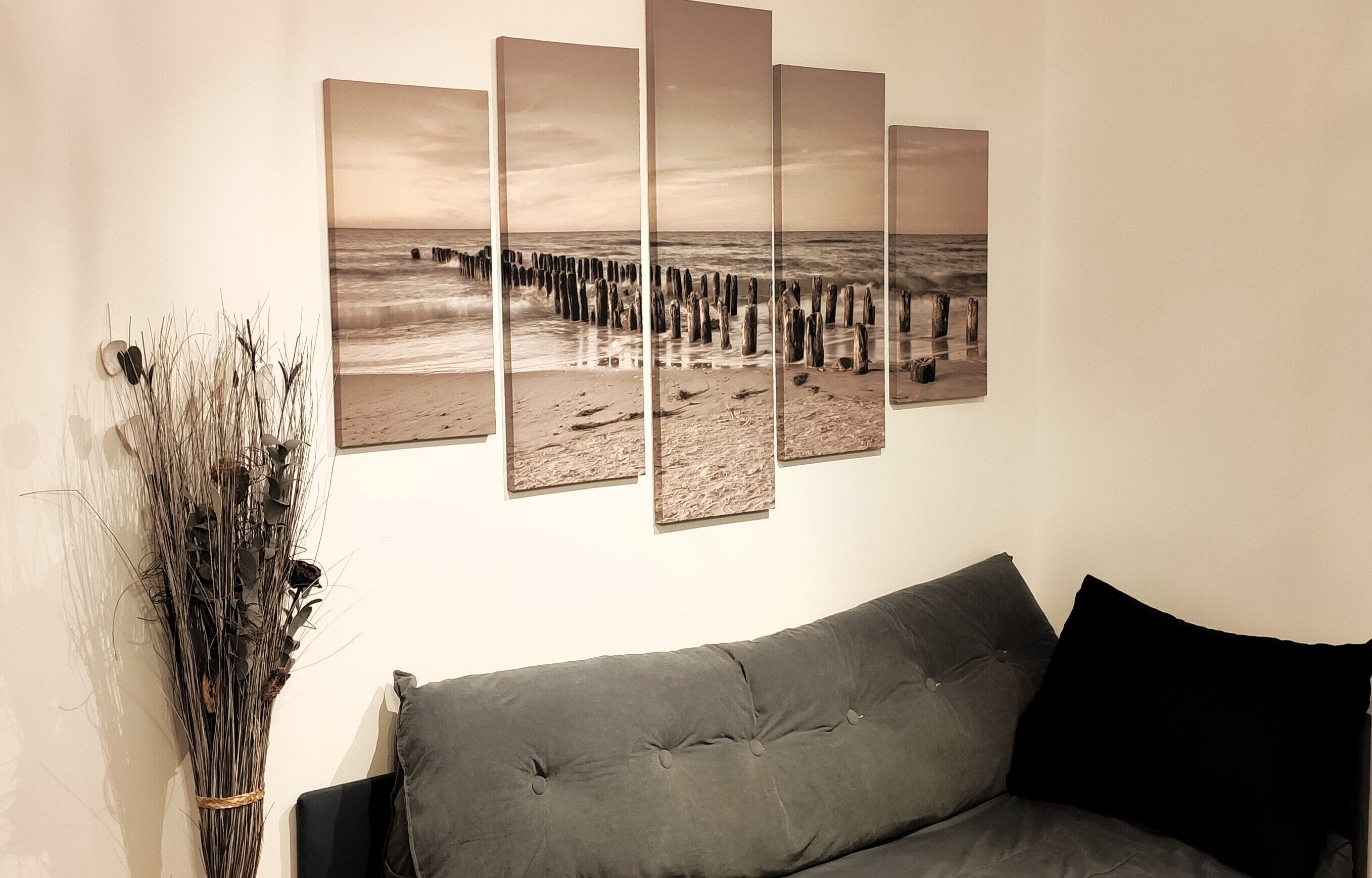 A room with a grey sofa, a wall-mounted artwork depicting a beach scene, and a tall vase of dried plants.