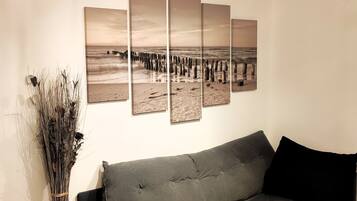 A room with a grey sofa, a wall-mounted artwork depicting a beach scene, and a tall vase of dried plants.