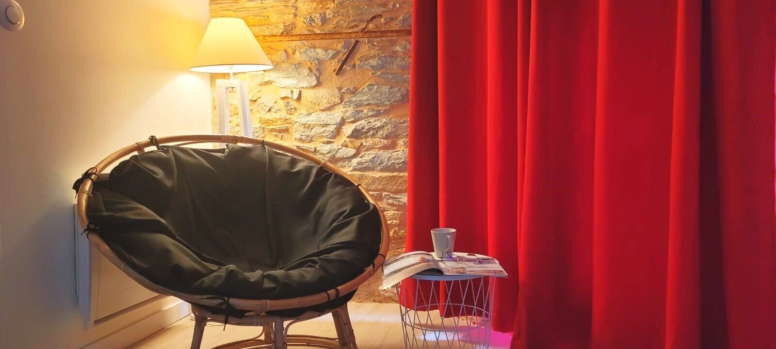 A room with a wicker chair, a lamp, a small table with a magazine, and a red curtain.