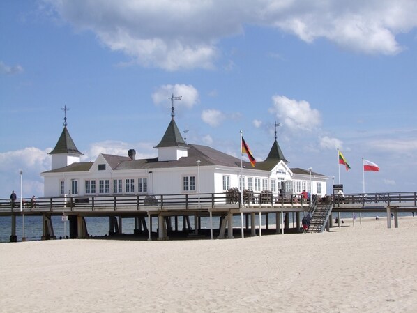 A white building with two green-roofed towers, a pier extending into the water, and flags on poles.