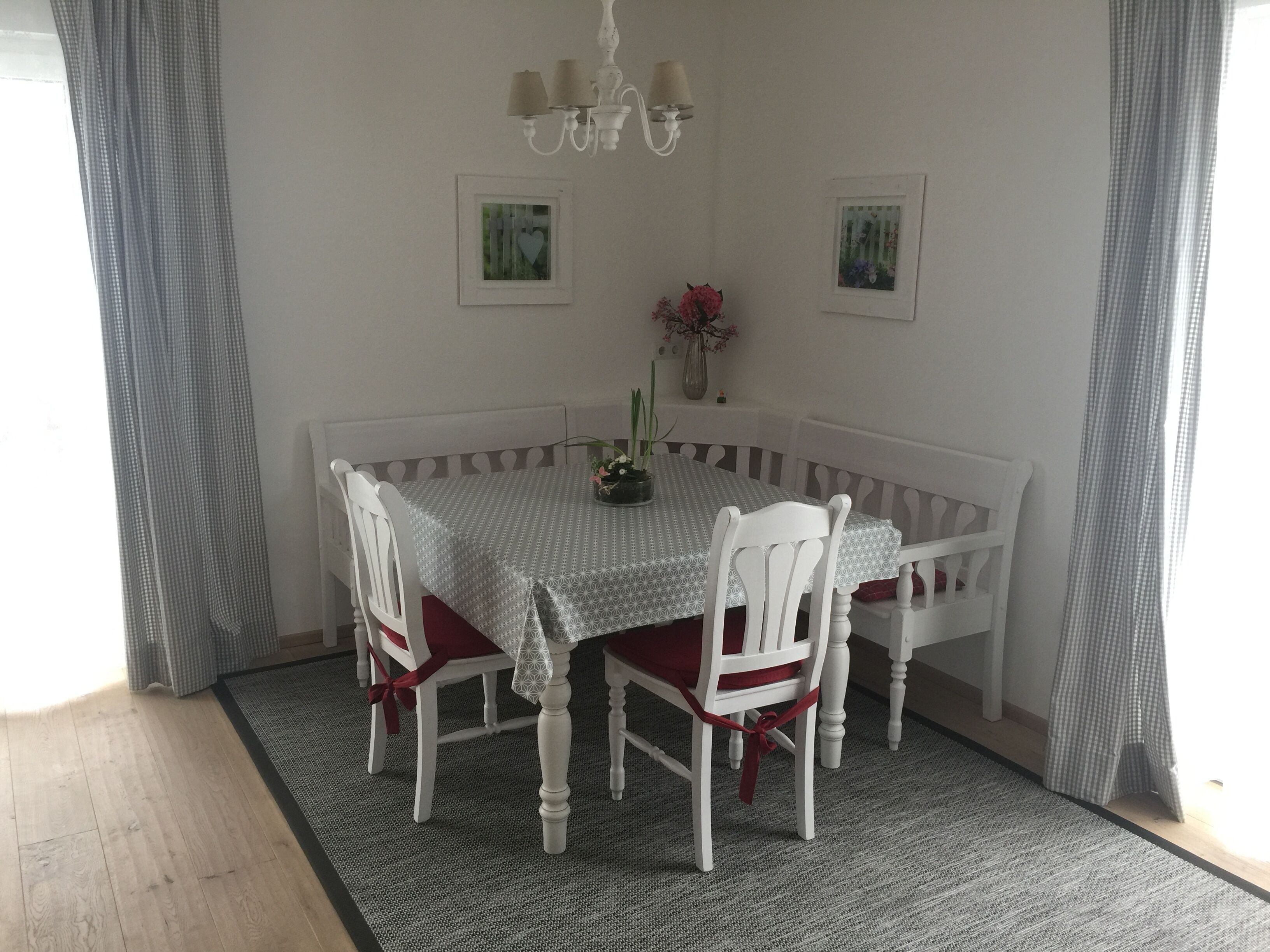 A dining table with a patterned tablecloth, white chairs with red cushions, a chandelier, and framed pictures on the wall.