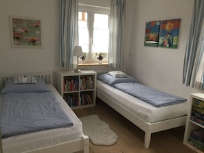 Una habitación con dos camas individuales, una estantería, una mesita con lámpara y un ventanal con cortinas.