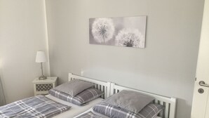 A bedroom with two beds, each with a white headboard and grey plaid bedding. There is a bedside table with a lamp and a small decorative item. A wall-mounted artwork of dandelions is visible.
