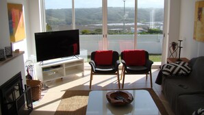 Un salon équipé d’un téléviseur, de deux fauteuils, d’un canapé et offrant une vue sur un pont et des collines.