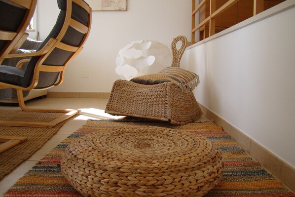 Una habitación con un tapiz tejido, un otomano redondo tejido, un pouf tejido y una silla de madera.