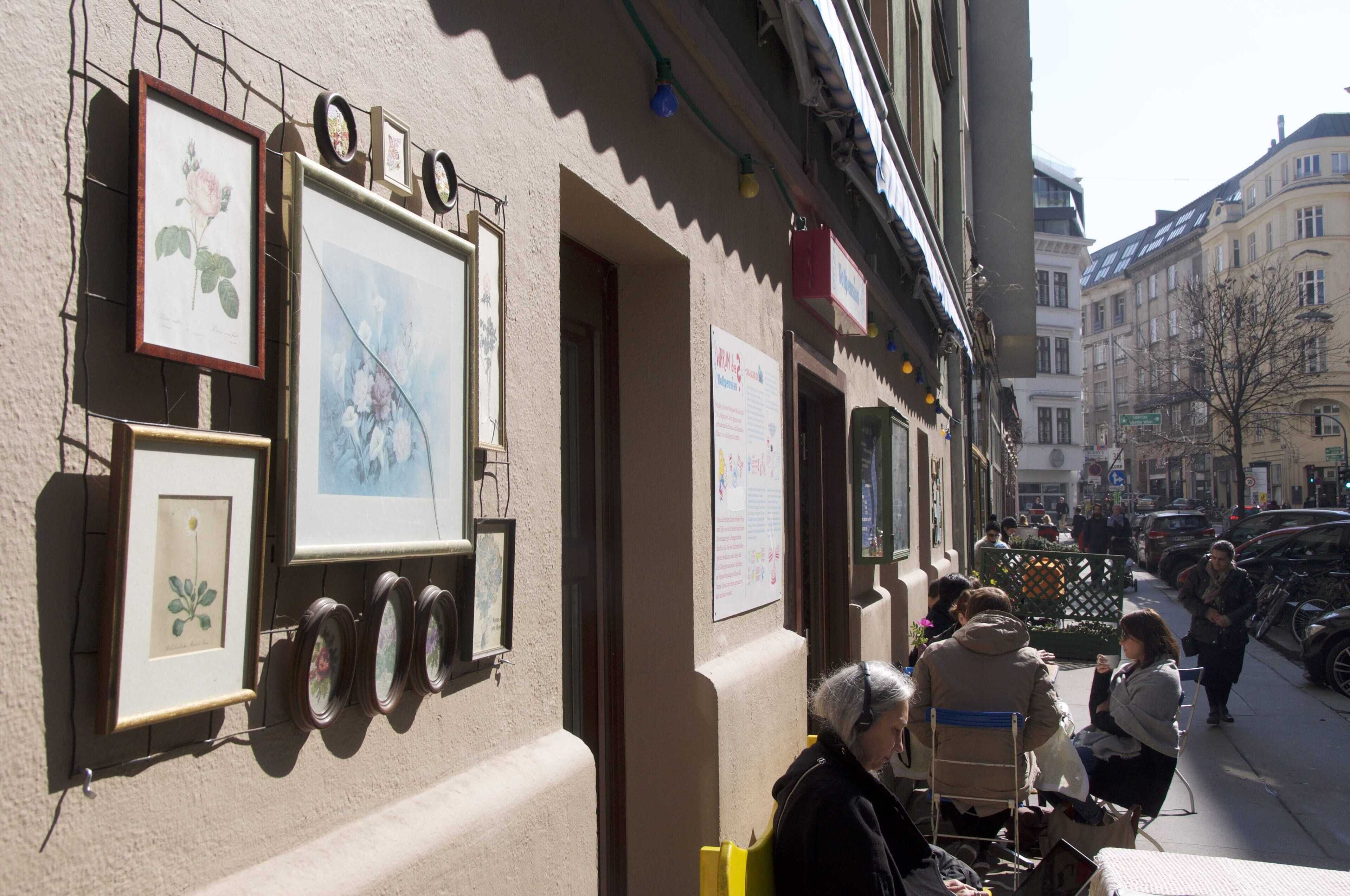 A building with framed botanical prints on the wall, a street with parked cars, and people sitting outside.