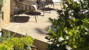 Un espace extérieur aménagé, abrité, avec des chaises en osier et une table, un luminaire suspendu et une vue sur la rue à travers une porte vitrée.