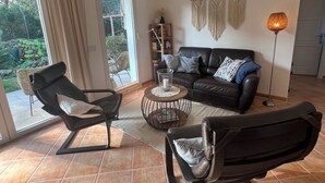 Un salon avec un canapé en cuir noir, deux fauteuils noirs, une table basse et un lampadaire. On y trouve également une bibliothèque et une grande fenêtre avec des rideaux.