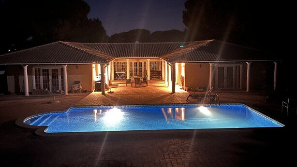 Une piscine avec une terrasse carrelée, un patio couvert doté de chaises et de tables, et un bâtiment avec un toit et des colonnes recouverts de carreaux.