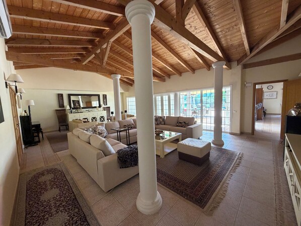 A spacious living area with high ceilings and wooden beams, featuring a sofa, coffee table, and a dining table.