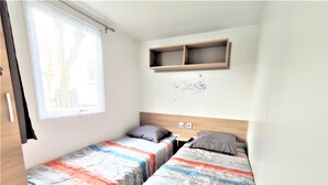 A small, well-lit room with two single beds, a wooden shelf above, and a window with blinds.