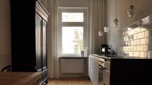 Une cuisine étroite avec un plancher en bois, une table à manger en bois, un meuble noir, une fenêtre avec des rideaux et un réfrigérateur en acier inoxydable.