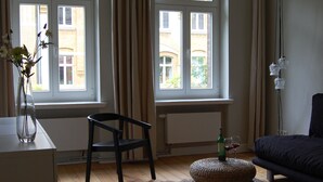 Une pièce avec deux fenêtres, une chaise, un canapé, une lampe et un vase rempli de fleurs.