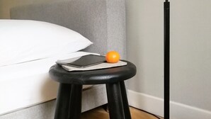 Un lit avec une tête de lit grise, un lampadaire noir, un petit tabouret sur lequel sont posés un livre et une orange, et un oreiller blanc.