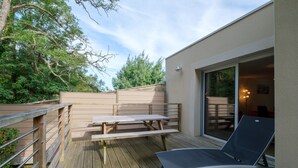 An outdoor deck with a wooden table and chairs, a lounge chair, and a glass door leading to an interior space.