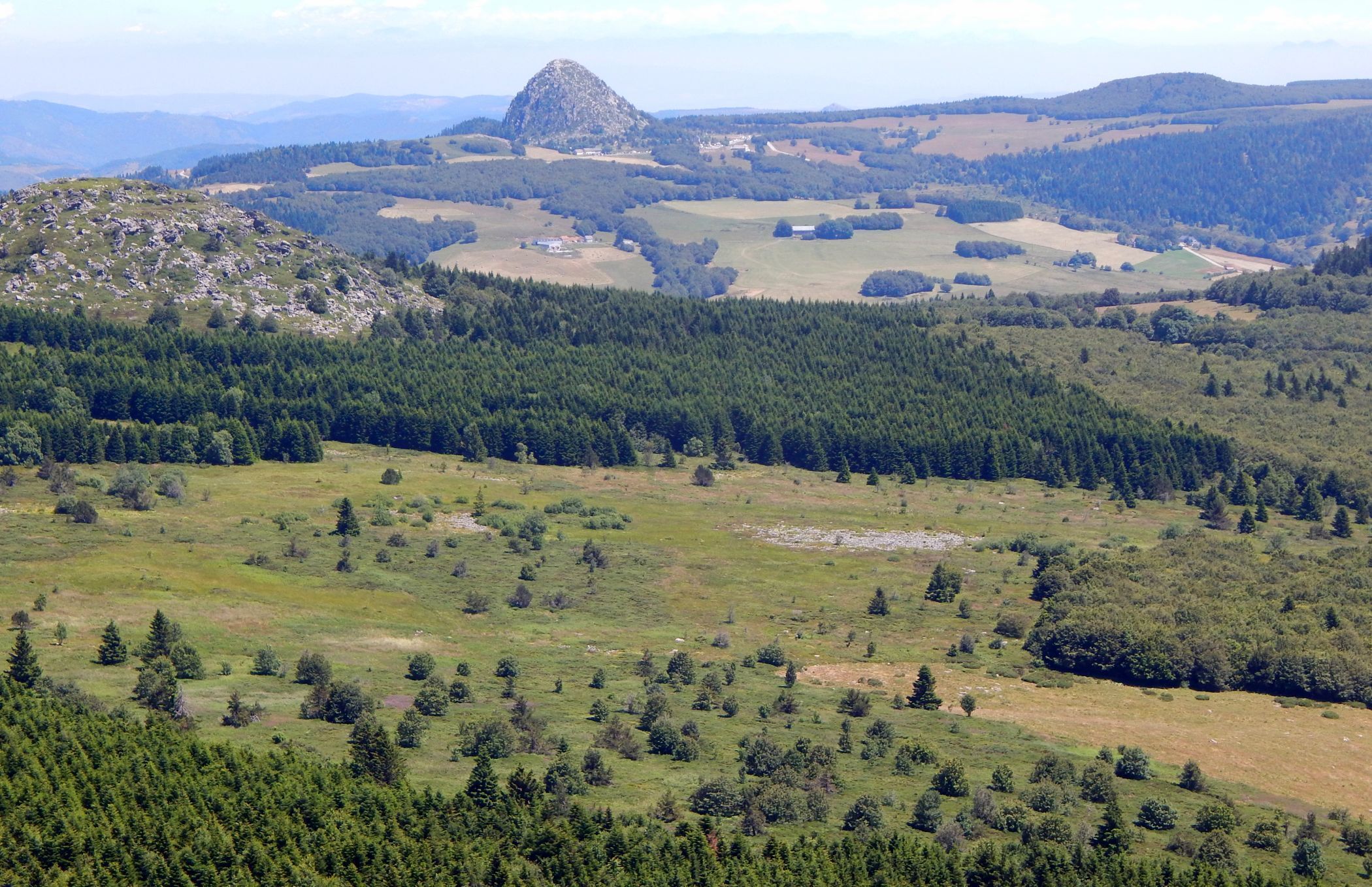 A panoramic view of a mountainous landscape with dense forests, open fields, and a prominent rocky hill.