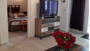 Quarto com cama, TV sobre um móvel de madeira e um vaso de flores vermelhas sobre uma mesa.